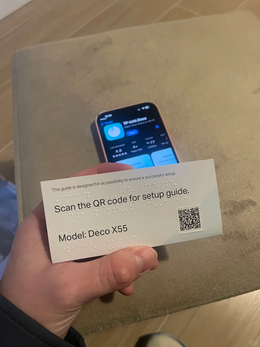 A hand holds an accessibility setup card with embossed Braille dots, an printed English guide reading 'This guide is designed for accessibility to ensure a successful setup. Scan the QR code for setup guide. Model: Deco X55', and a QR code, with a smartphone in the background displaying the TP-Link Deco app page on an App Store.