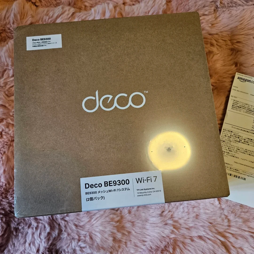 Cardboard packaging box for a Deco BE9300 Wi-Fi 7 mesh system resting on a pink fuzzy surface next to an Amazon packing slip.