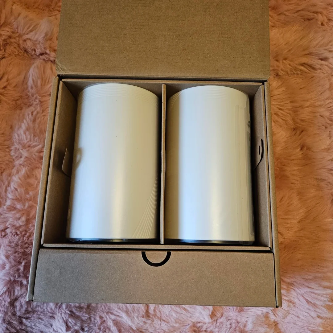 Two white cylindrical mesh Wi-Fi routers packaged side by side in an open cardboard shipping box resting on a pink furry blanket.