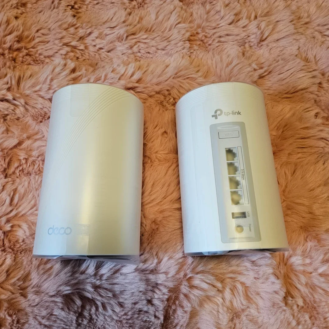 Two white TP-Link Deco cylindrical mesh Wi-Fi units wrapped in clear protective film resting on a fluffy pink fur surface. The left unit shows the front with the Deco logo, while the right unit shows the rear panel with Ethernet ports, a USB port, and a power connection.