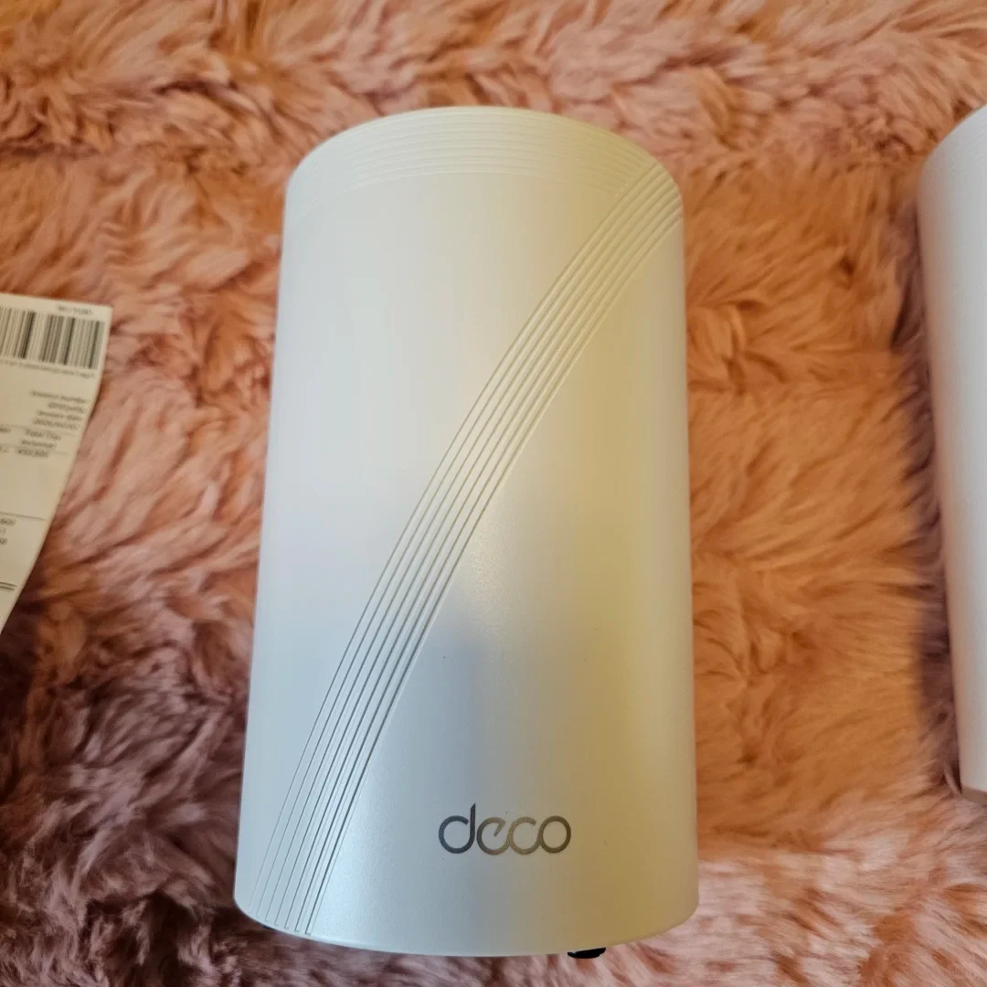 A white cylindrical TP-Link Deco mesh Wi-Fi unit with diagonal grooved lines standing on a pink shaggy rug next to a paper receipt.