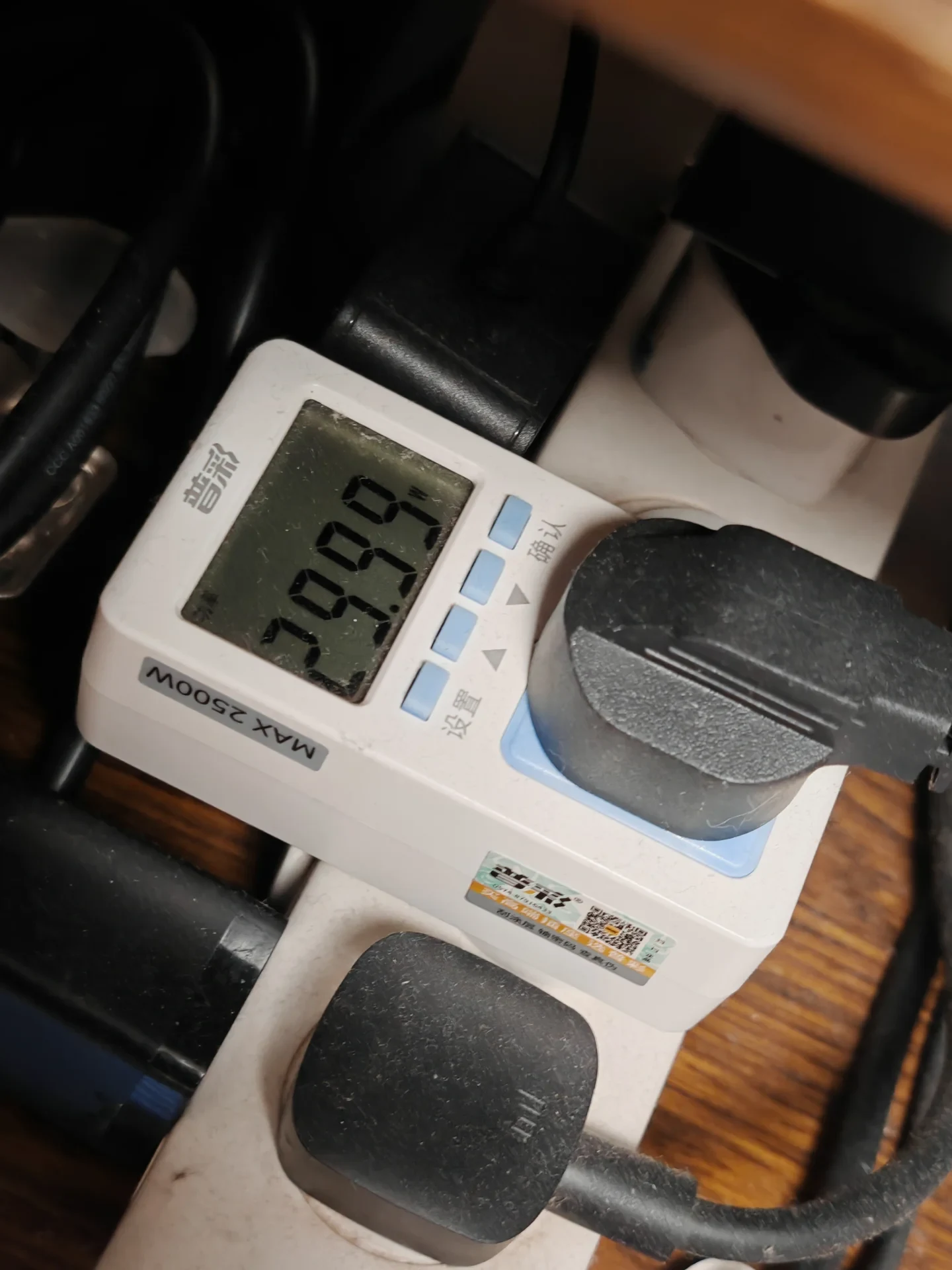 A white plug-in digital power meter plugged into an extension cord, displaying a power reading on its LCD screen with a black electrical plug connected.