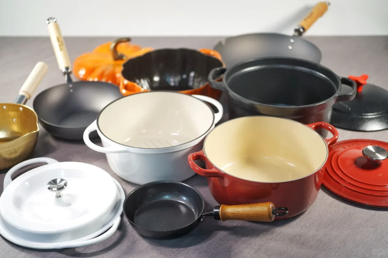 An assortment of cookware arranged on a surface, including enameled cast iron Dutch ovens in white, red, black, and an orange pumpkin shape, as well as a small skillet, woks with wooden handles, and a brass saucepan.