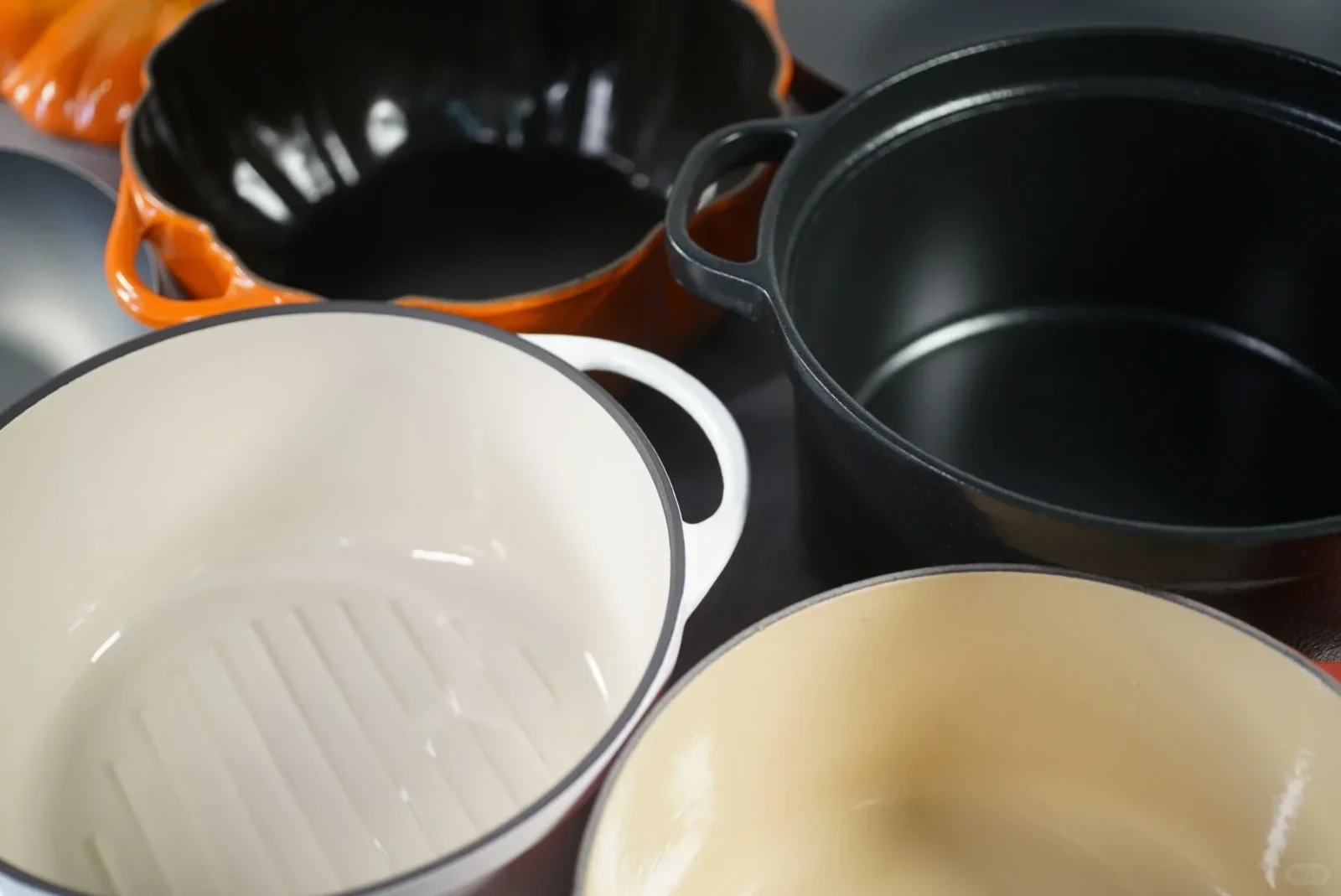 The interiors of four enameled cast-iron Dutch ovens viewed from above: a pale Vermicular pot, cream-colored Le Creuset, black Ito pot and fluted black Staub Pumpkin.