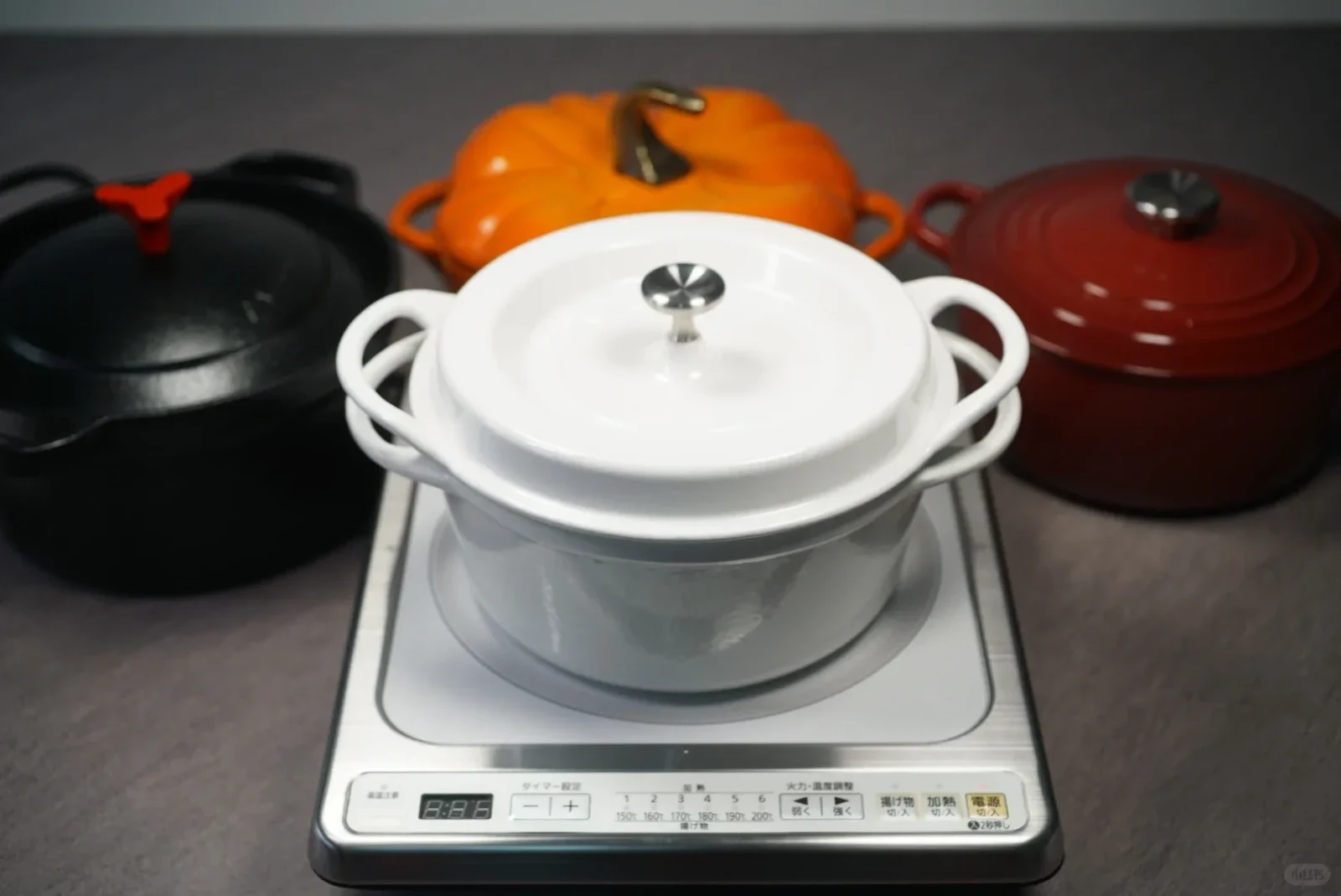 A white enameled cast iron Dutch oven sits on a silver countertop induction cooktop, surrounded by a black pot, an orange pumpkin-shaped cocotte, and a red Dutch oven.