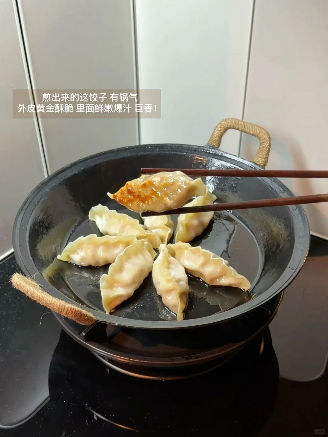 Dumplings pan-frying in a black pan on a stovetop, with chopsticks holding up one dumpling to display its golden-brown, crispy bottom.
