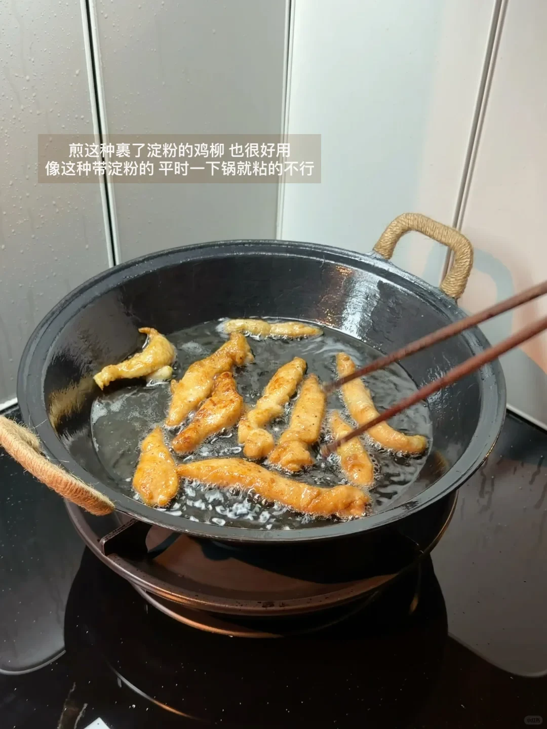 Battered chicken strips frying in oil inside a black wok on a stovetop, held and flipped with wooden chopsticks.
