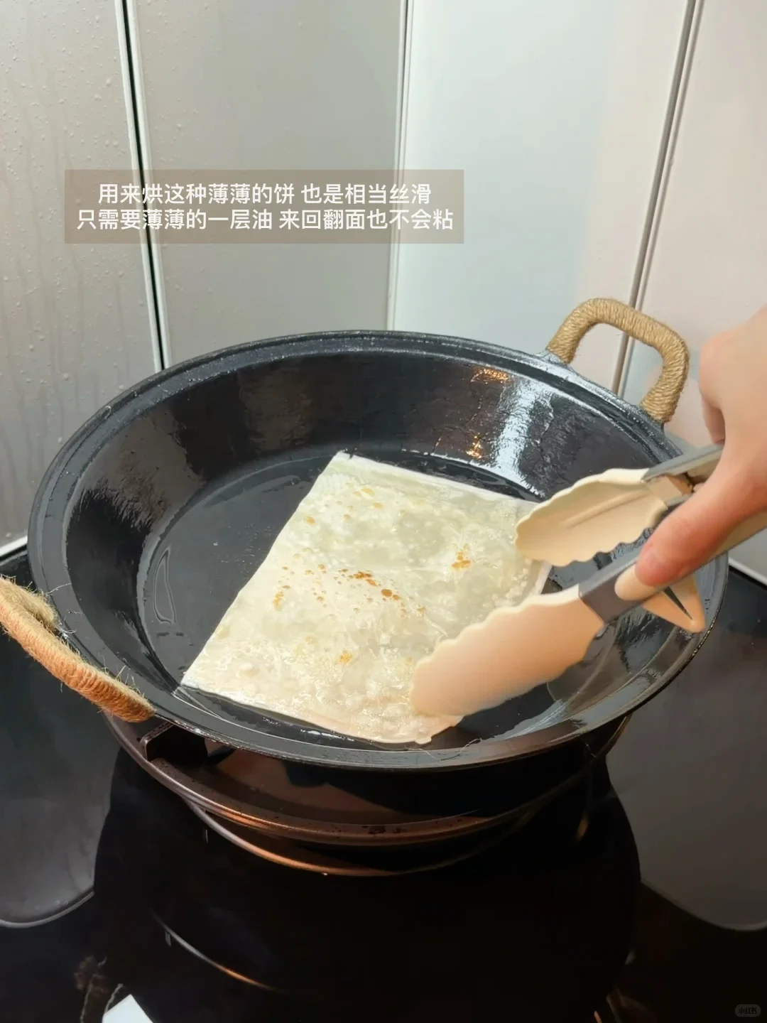 A square flatbread being flipped with tongs in an oiled black iron pan on a stovetop.