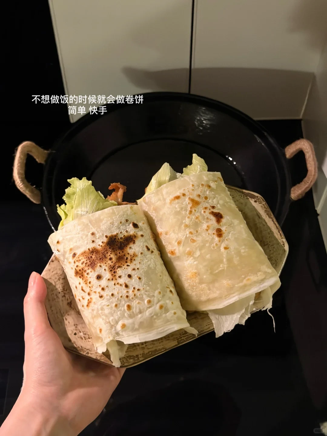 A hand holding a plate with two cooked wraps filled with lettuce and meat in front of a black skillet on a stovetop.