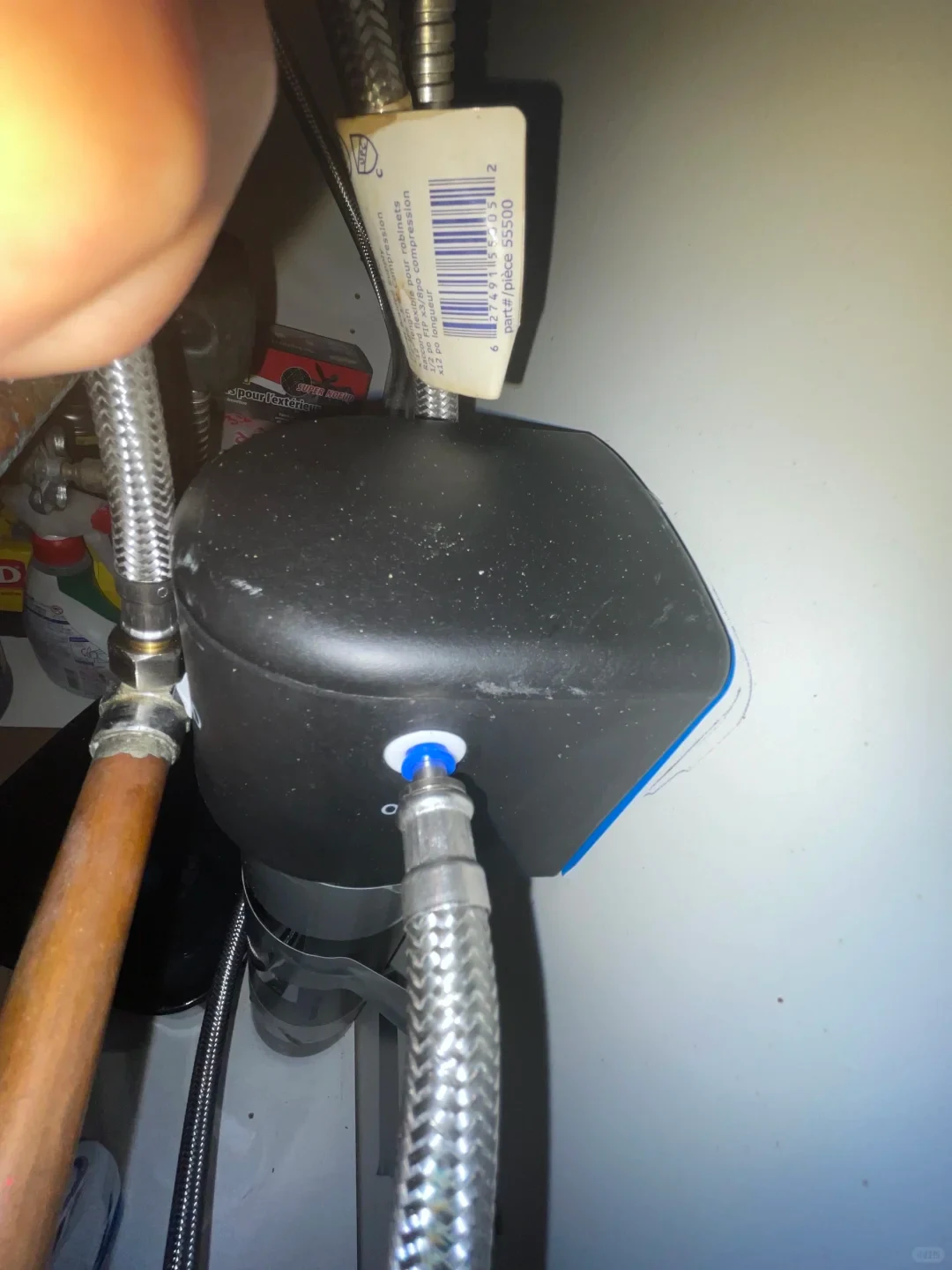 A close view of the black filter head beneath the sink, with braided metal hoses attached and a copper pipe nearby. A hose tag with a barcode hangs above it.
