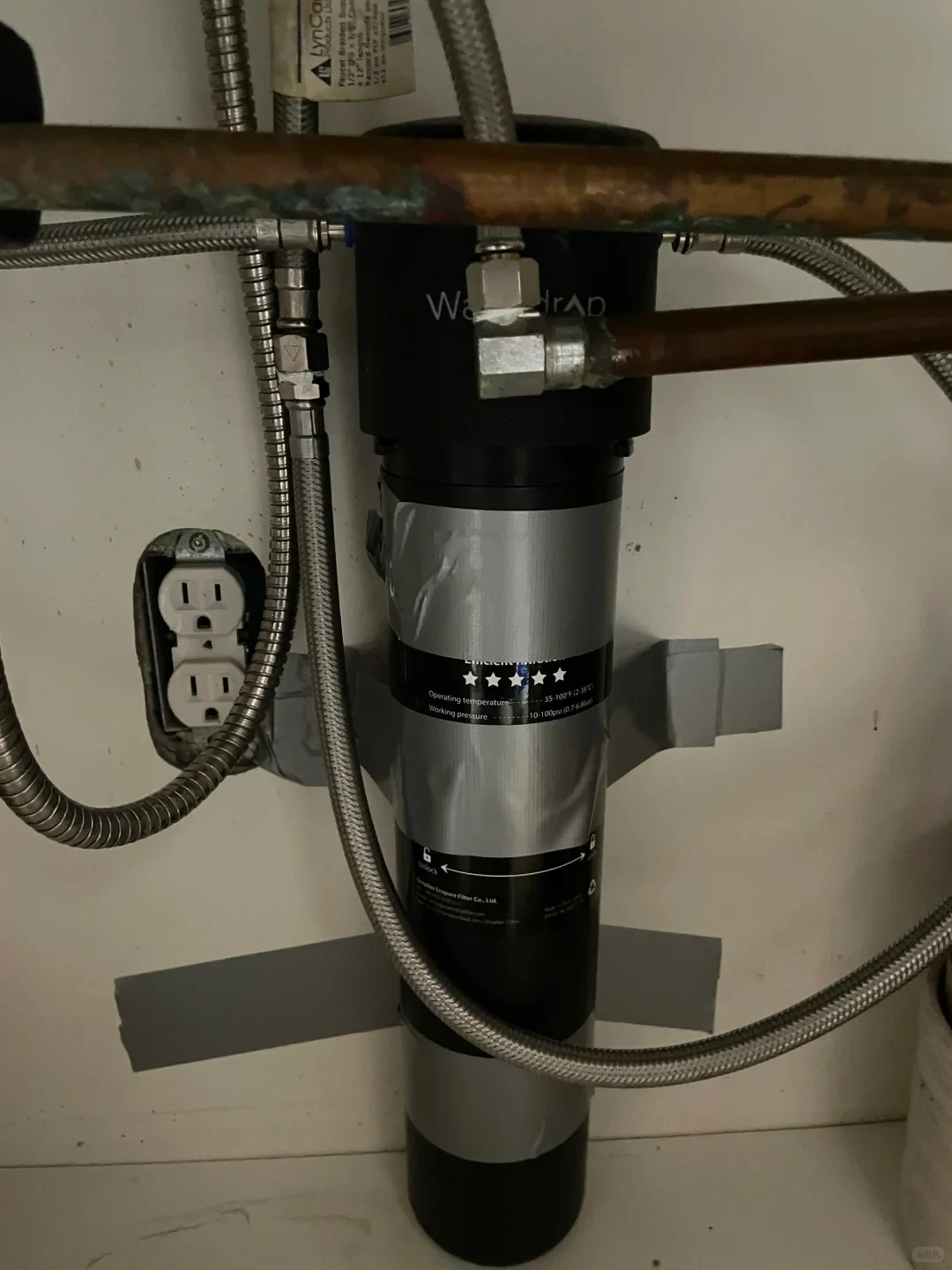 A vertically mounted cylindrical water filtration unit beneath a sink, connected to copper pipes and braided hoses beside an electrical outlet.