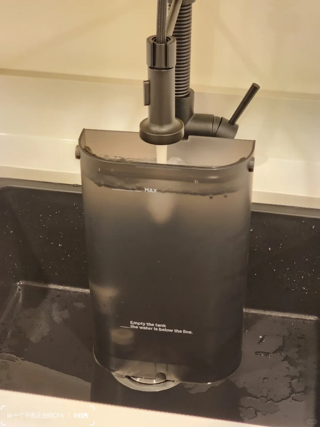 A dark translucent water tank is positioned beneath a running kitchen faucet over a wet sink. Water reaches the “MAX” mark, and the tank says to empty it when the water is below the line.