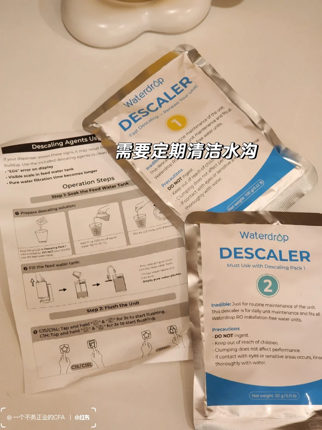 Two matching Waterdrop descaler sachets, numbered 1 and 2, lie over a partly obscured instruction sheet with feed-tank soaking and unit-flushing stages. Packet 1 is marked 100 g; packet 2 is marked 50 g and says to use it with packet 1. A Chinese callout calls for regular cleaning of the water passages.