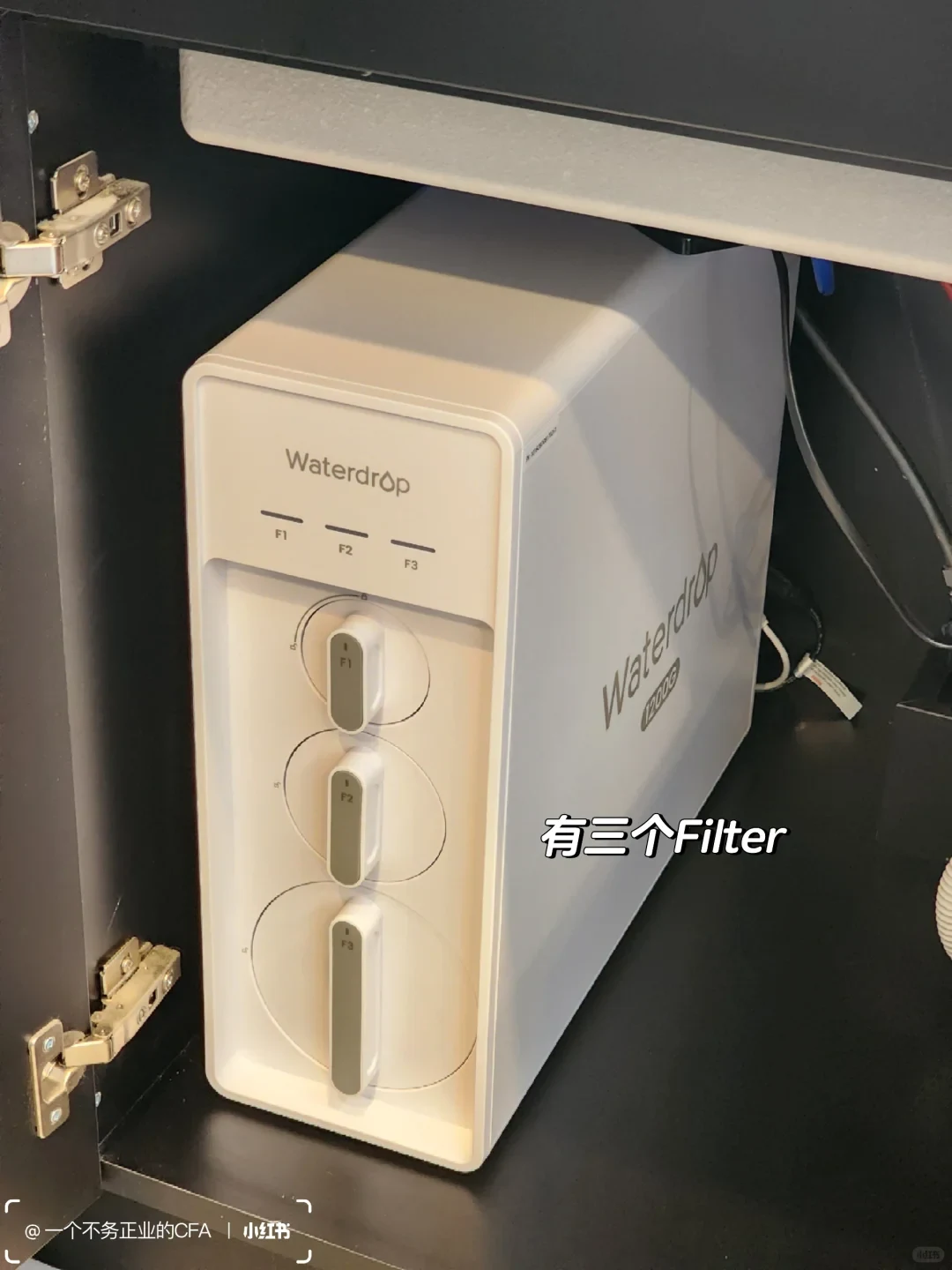 A white Waterdrop water filtration unit is installed inside a dark under-sink cabinet, with three vertically arranged filter cartridges labeled F1, F2, and F3. An overlaid caption says it has three filters.