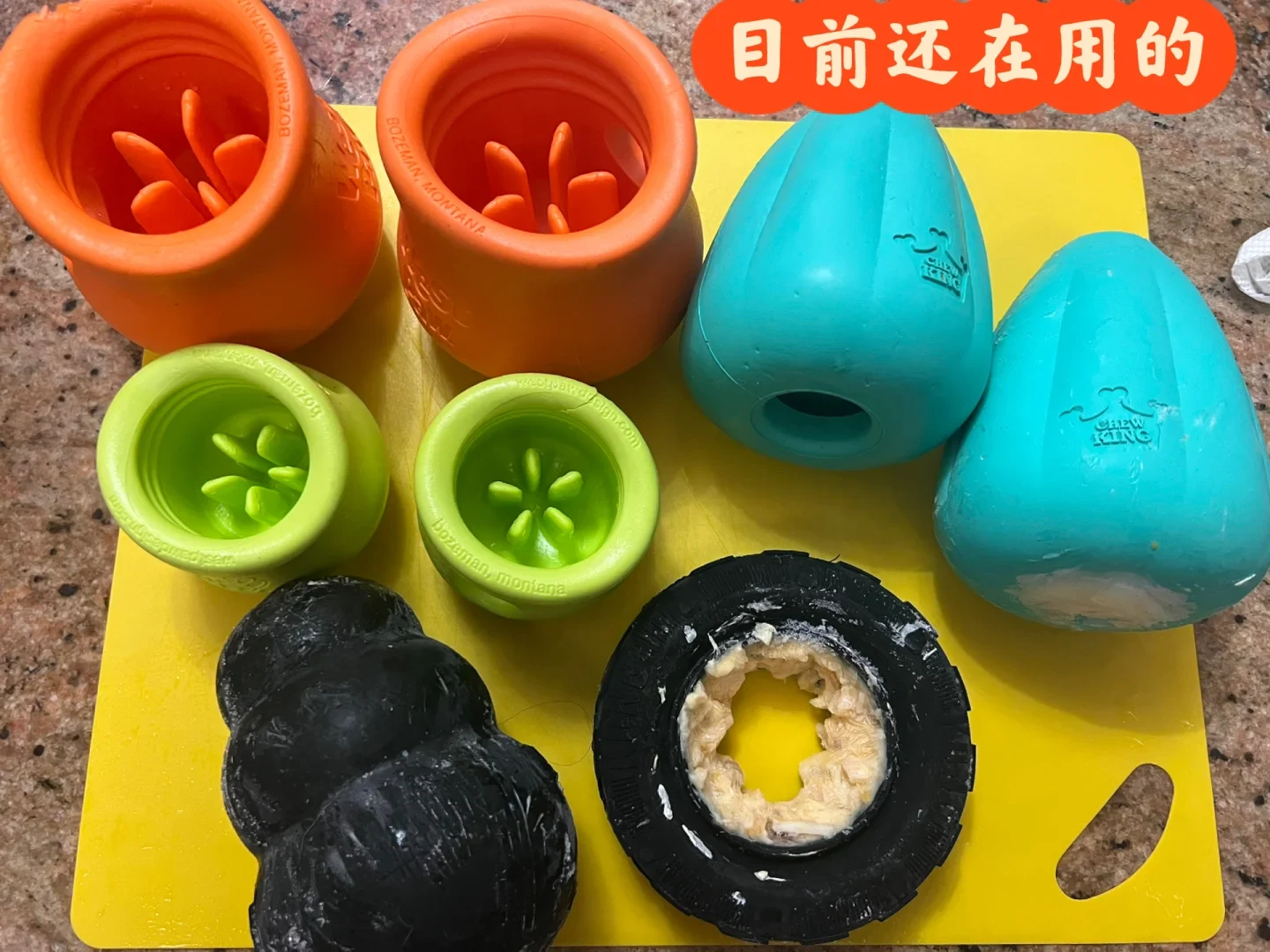 An assortment of rubber dog enrichment toys arranged on a yellow mat, including orange and lime green West Paw treat toys, two teal Chew King toys, a black Kong toy, and a black tire toy stuffed with paste.