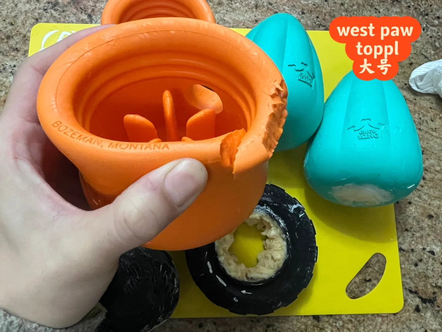 A hand holds a chewed and damaged bright orange West Paw Toppl dog toy above a yellow cutting board, alongside two teal Chew King toys and black rubber enrichment toys filled with food.