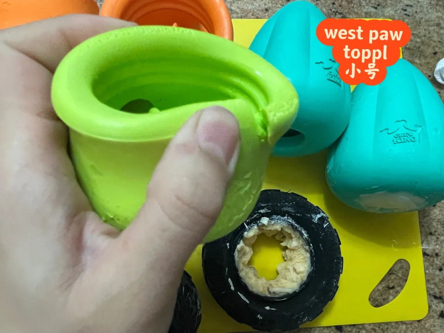 A hand holds the damaged rim of a green small West Paw Toppl beside other food-stuffing toys.