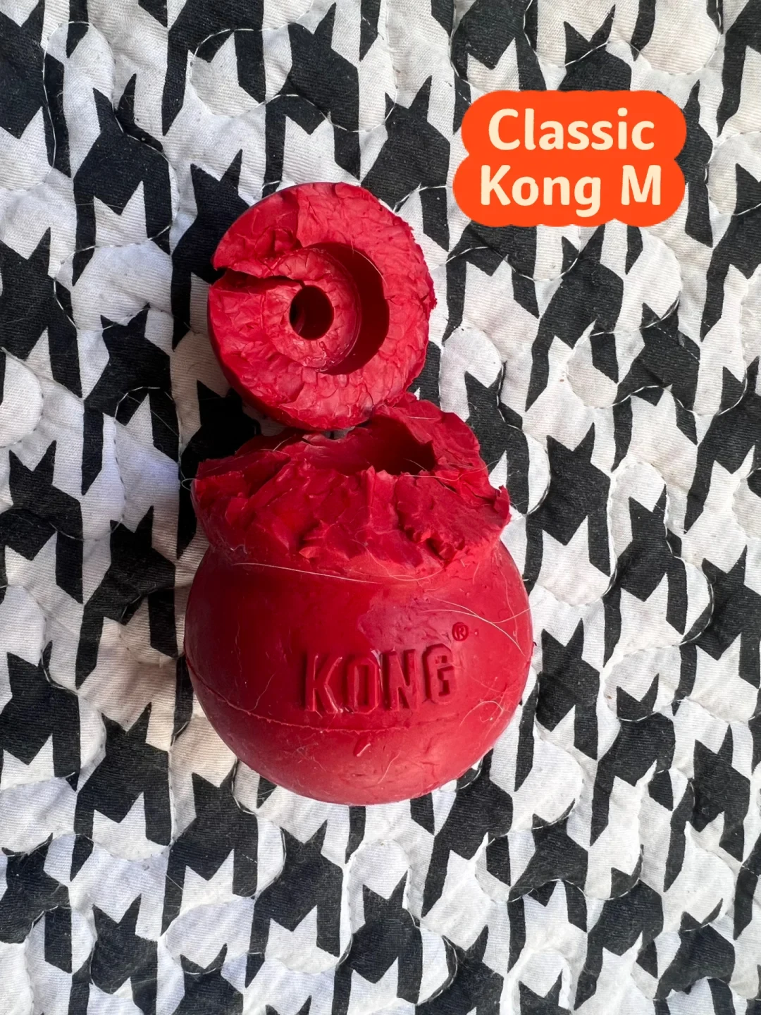 A chewed and broken red rubber KONG Classic dog toy split into two pieces, resting on a black-and-white houndstooth patterned blanket with an orange label reading 'Classic Kong M'.