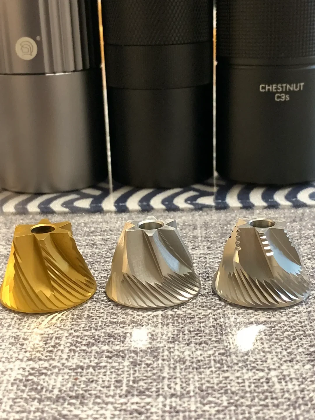 Three metal coffee-grinder burrs with spiral cutting teeth—one gold-colored and two silver—are arranged on a textured surface in front of black grinder containers.