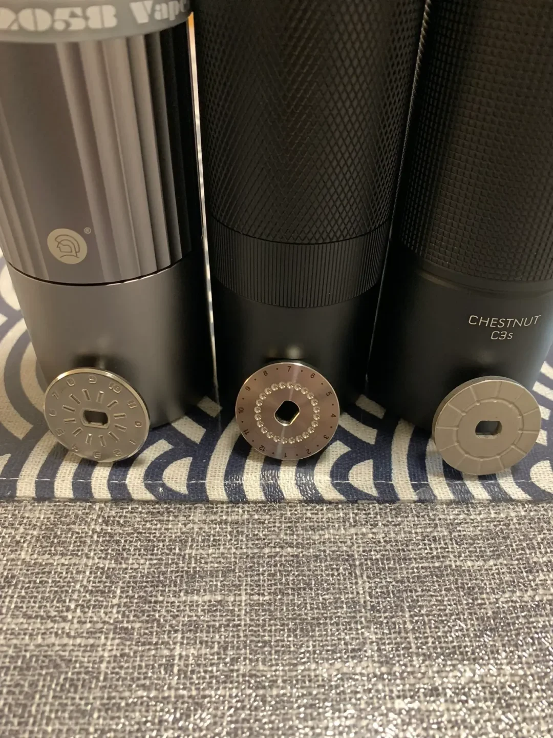 Close-up of three dark cylindrical coffee grinders standing side by side, each with a round adjustment dial or cap placed in front. The right grinder is labeled “CHESTNUT C3S.”