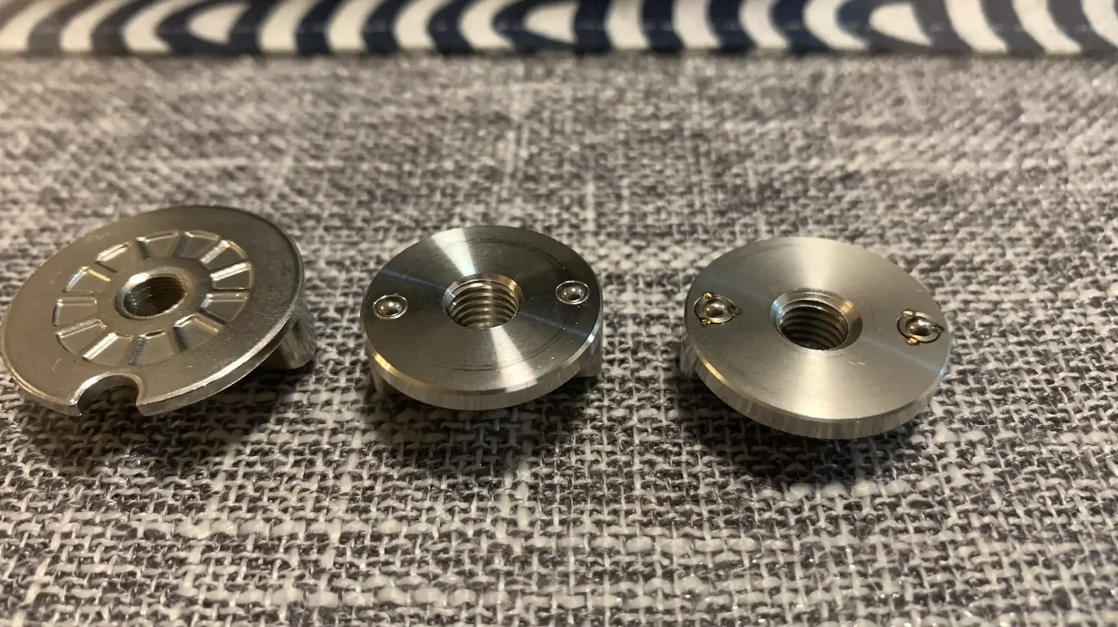 Three small round machined metal parts with central threaded holes and recessed details lie on a woven gray fabric surface.