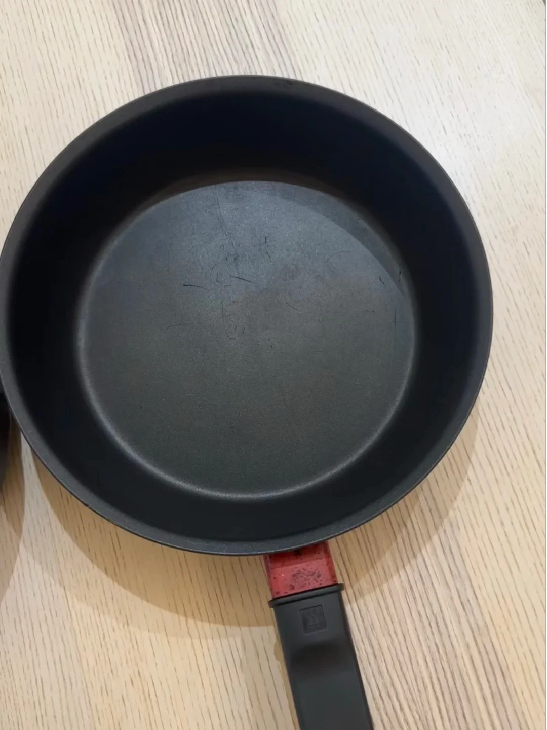 Top-down view of a black Zwilling non-stick frying pan with surface scratches on its interior coating, placed on a light wooden surface.