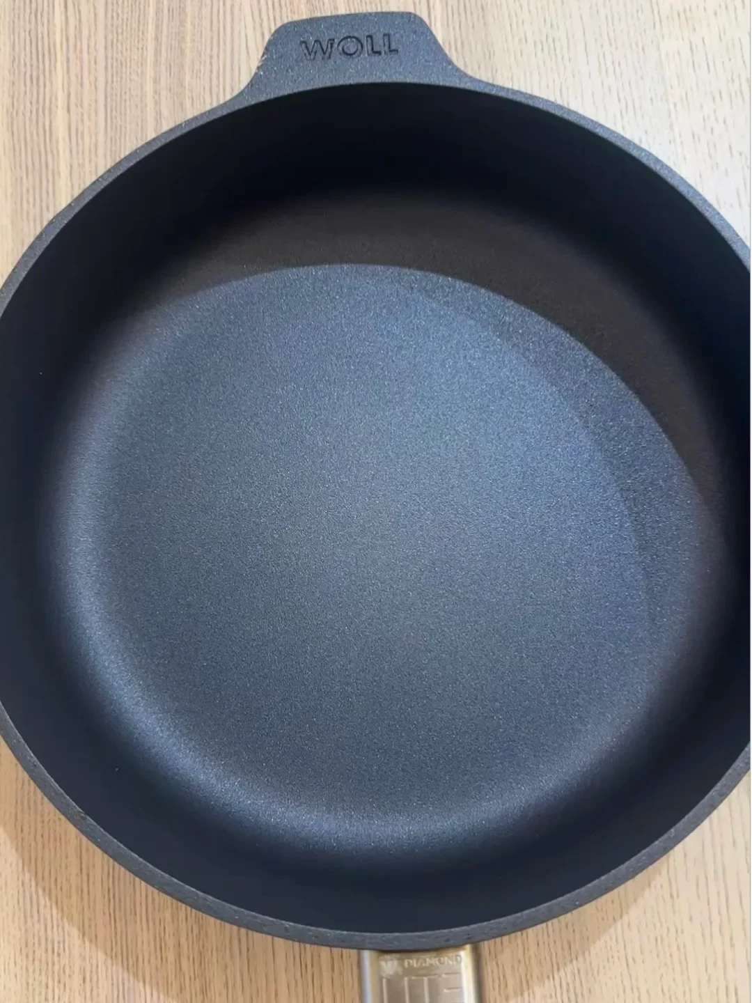 Top-down view of a black Woll nonstick frying pan with an intact cooking surface resting on a wooden tabletop.