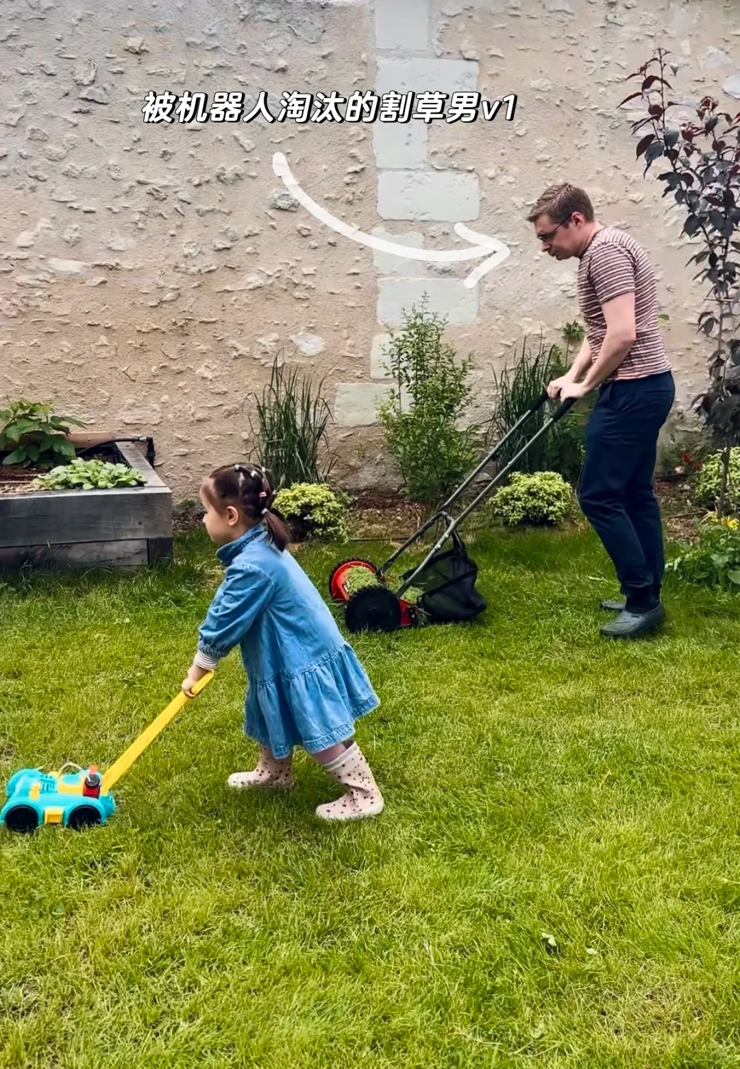 A young girl in a blue dress pushes a toy lawn mower across a grassy yard while a man behind her mows the lawn with a manual push mower.
