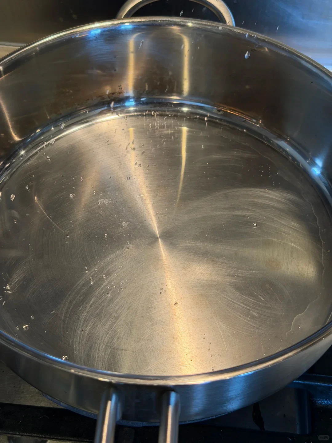 Close-up overhead view of an empty stainless steel pan on a stovetop with light swirl marks and reflections on the bottom surface.