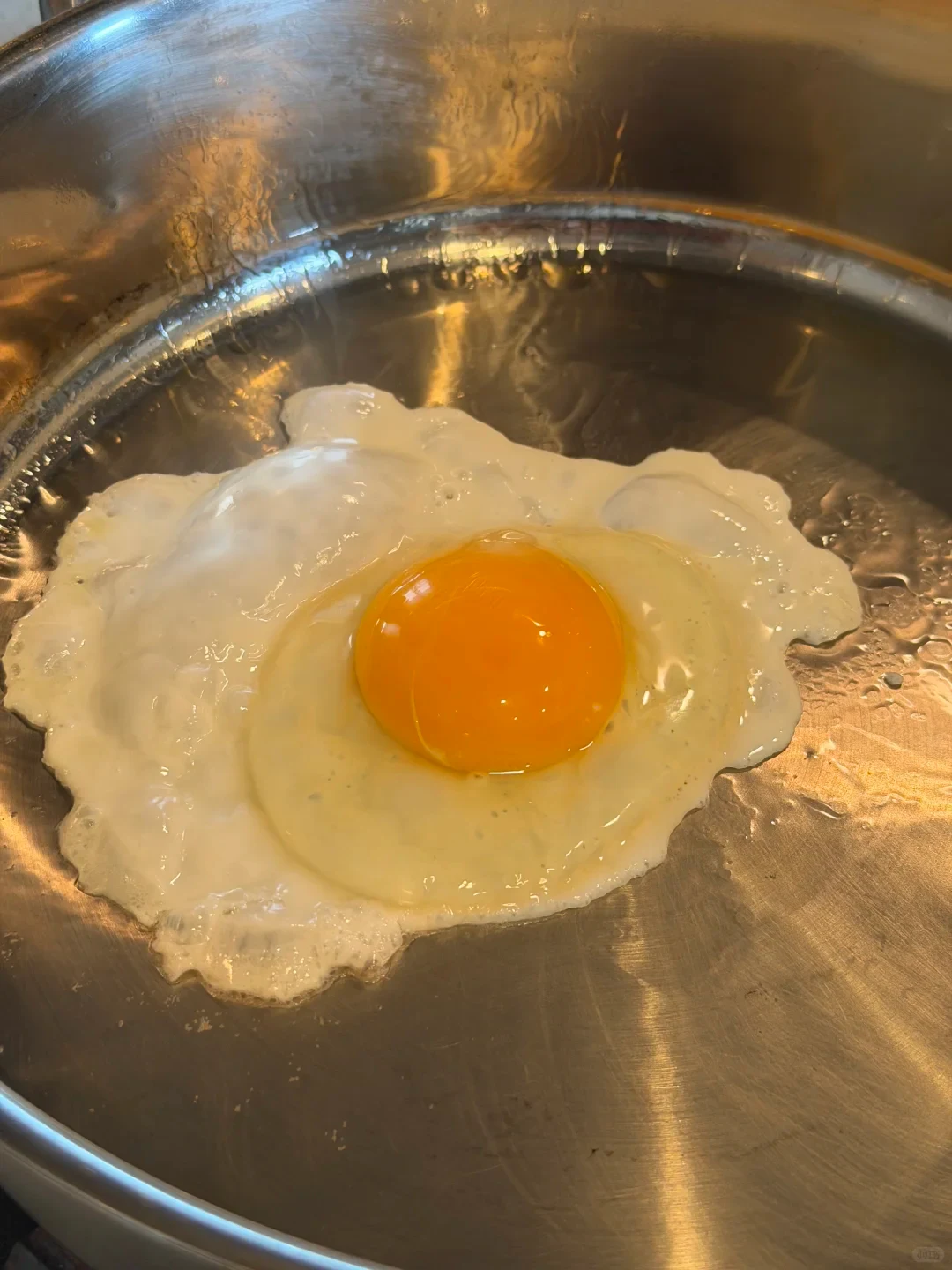 A sunny-side-up egg with an intact, bright yellow-orange yolk cooking in a stainless steel frying pan.