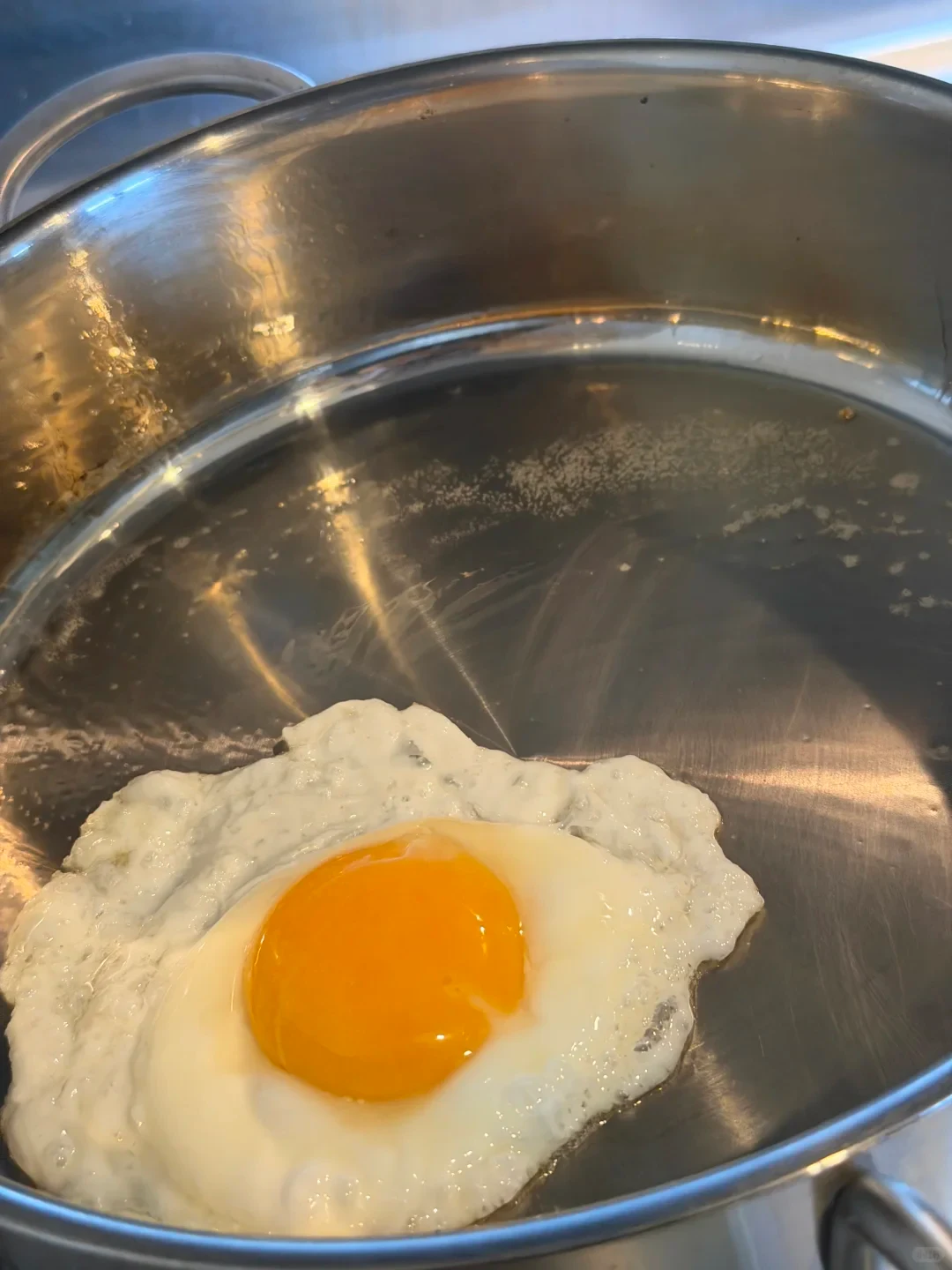 A sunny-side-up fried egg with a bright yellow yolk cooking inside a stainless steel pan.