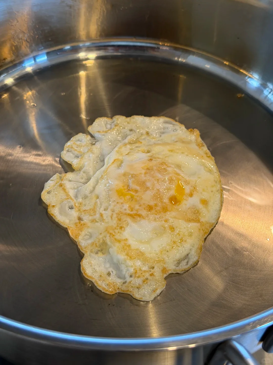 A fried egg with lightly browned edges cooking in the center of a stainless steel skillet.